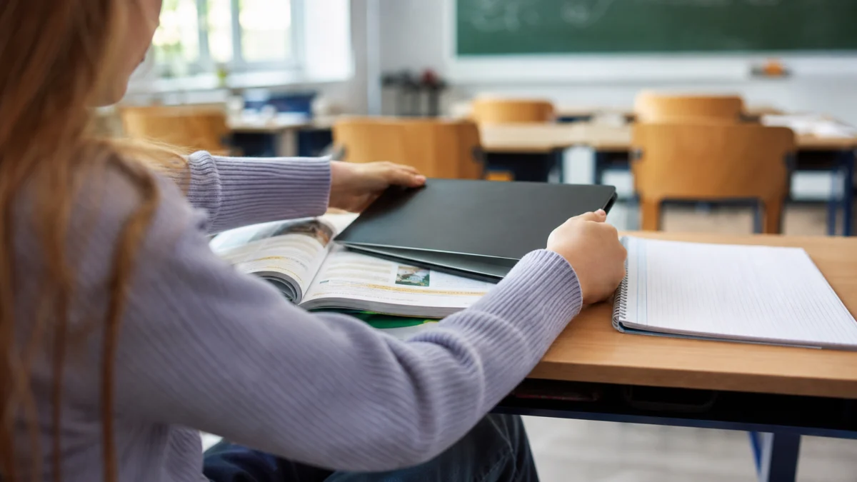 Symbolbild: Schülerin schließt im Klassenraum ein Tablet und wendet sich Schulbuch und Heft zu